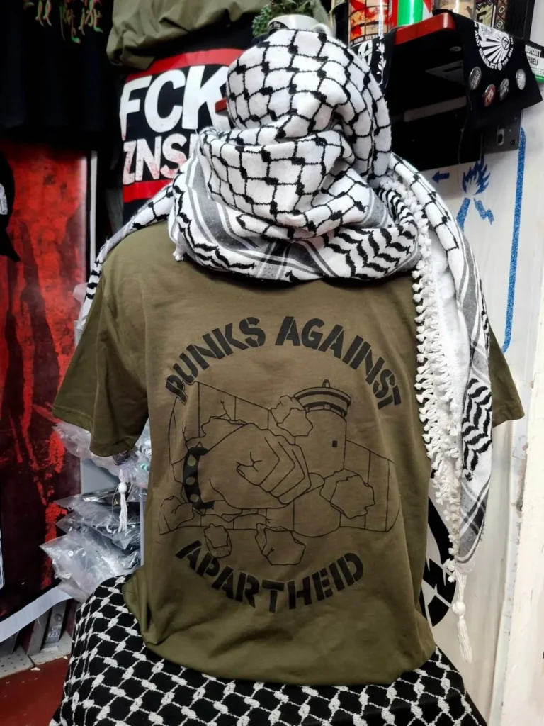 Slide 1: A headless mannequin wearing a kuffiyyeh on top and a green T-shirt with black stencil PAA logo, which shows the apartheid wall being smashed through by a fist with a spikey bracelet and the words PUNKS AGAINST APARTHEID around the circle’s edge.