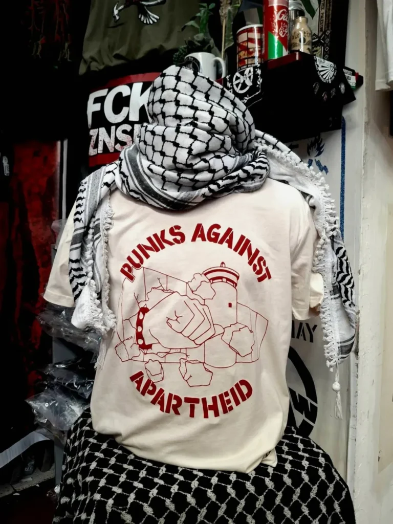 Slide 2: A headless mannequin wearing a kuffiyyeh on top and a light beige T-shirt with dark red stencil PAA logo, which shows the apartheid wall being smashed through by a fist with a spikey bracelet and the words PUNKS AGAINST APARTHEID around the circle’s edge.