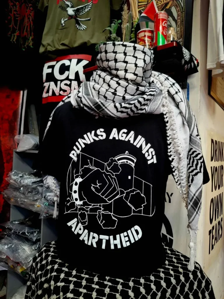 Slide 3: A headless mannequin wearing a kuffiyyeh on top and a black T-shirt with white stencil PAA logo, which shows the apartheid wall being smashed through by a fist with a spikey bracelet and the words PUNKS AGAINST APARTHEID around the circle’s edge.