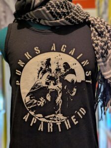 Slide 4: A headless mannequin wearing a kuffiyyeh on top and a black T-shirt with white stencil logo, which shows a group of masked and kuffiyyeh wearing individuals gathered around and PUNKS AGAINST APARTHEID around the circle’s edge.