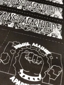 Slide 7: Another close-up shot of another half of patches, with a different alternate circular logo visible: the apartheid wall being broken through by a front-facing fist with spikes, and the text PUNKS AGAINST APARTHEID on the top and bottom.