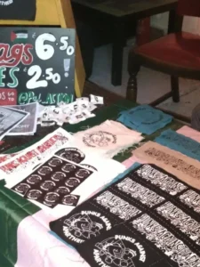 Slide 8: A table full of merch selling flags, badges, stickers, and patches at a punk show.