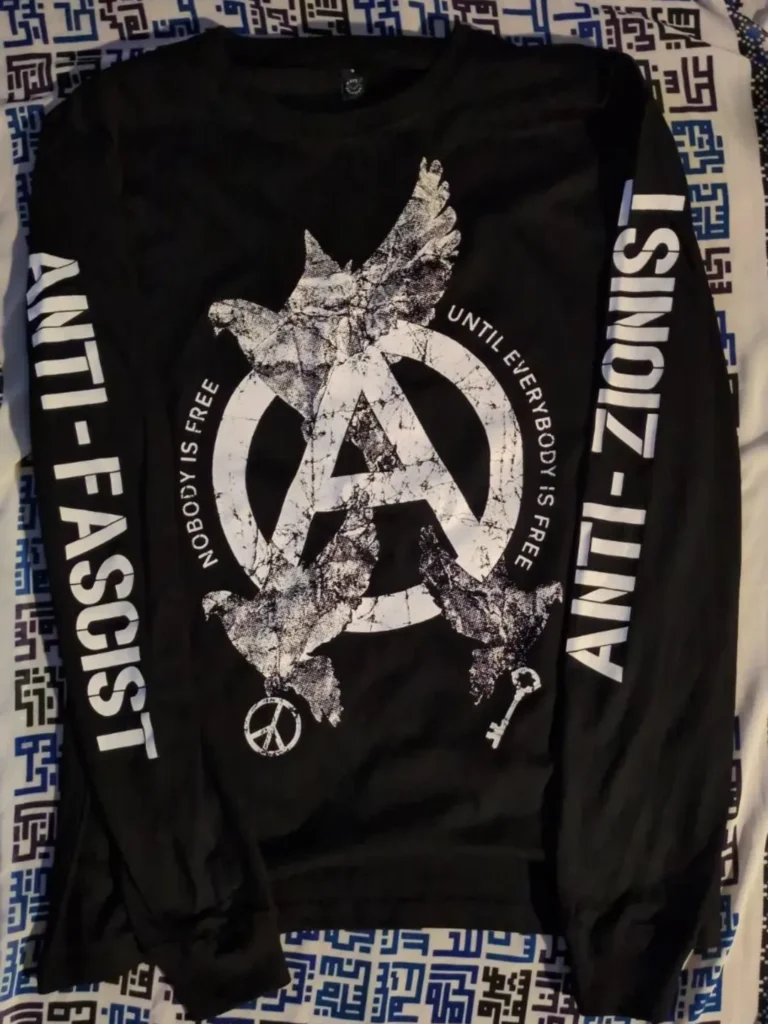 A long sleeve T-shirt on the floor on top of a large scarf with the names of Palestinian villages in Kufic Arabic style square calligraphy font.The shirt shows a large rough circle (A) with three large doves emerging in triangle formation from the tips of Thea, with the bottom two doves holding a peace sign and a large key on the left and right respectively. On the edge of the (A) it says NOBODY IS FREE//UNTIL EVERYBODY IS FREE on the left and right respectively. And on the left (the right arm), it says ANTI-FASCIST; on the right (the left arm) it says ANTI-ZIONIST.