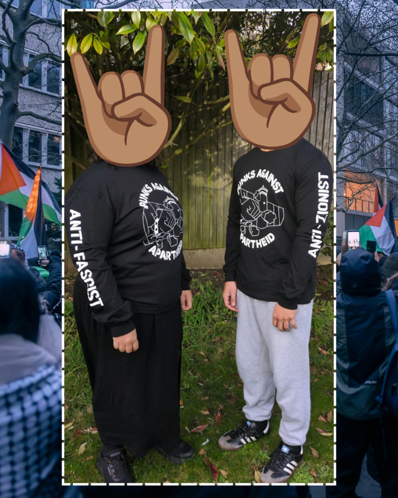 A background showing a Palestine protest. In the middle with a white outline, a photo of two individuals with a long-sleeve Punks Against Apartheid t-shirt on, standing facing each other in a garden and looking forwards. Their heads have been replaced by large 'metal hands' emojis. The centre of the shirt shows the PAA logo (a fist with a spikey bracelet punching through the apartheid wall), and each exposed arm shows the phrases: 'ANTi-ZIONIST' and 'ANTI-FASCIST'.