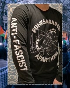 A photo of an individual facing forwards from the neck downards, showing the PAA logo (a fist with a spikey bracelet punching through the apartheid wall) & one arm which says ANTI-FASCIST.